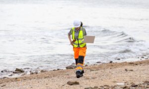 A land surveyor in safety gear using a laptop to record boundary survey data along a shoreline during an environmental cleanup